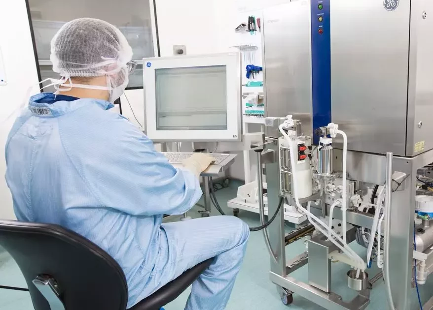 A scientist in full protective gear operates a large biopharma manufacturing machine, ensuring GMP standards and quality control via a computer interface.