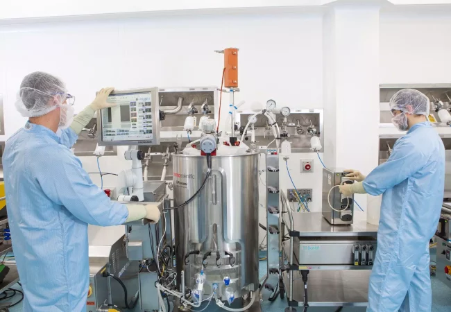 Two scientists in full protective gear manage a GMP (Good Manufacturing Practice) biomanufacturing process, monitoring a large stainless steel bioreactor. One operates a control panel while the other handles tubing connections, ensuring compliance for QP (Qualified Person) release. This process ensures product quality and safety before distribution.