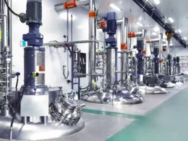 A row of large, industrial stainless-steel bioreactors with attached pipes and control units is shown in a brightly lit, clean laboratory or production facility, essential for bioanalytical development. The floor is green, the machines are labeled, and the background features more equipment and infrastructure.