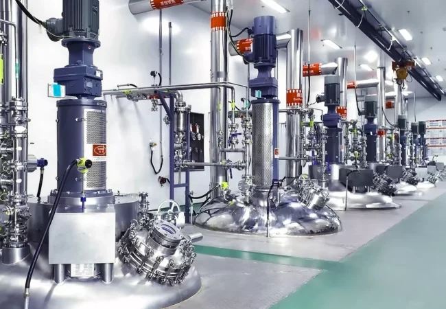 A row of large, industrial stainless-steel bioreactors with attached pipes and control units is shown in a brightly lit, clean laboratory or production facility, essential for bioanalytical development. The floor is green, the machines are labeled, and the background features more equipment and infrastructure.