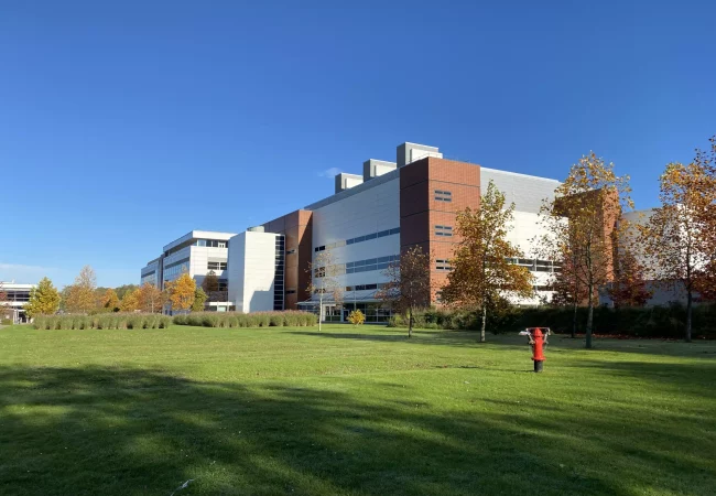 A modern building at the Hoddesdon site on a sunny day, surrounded by trees with autumn leaves and a grassy lawn with a red fire hydrant.
