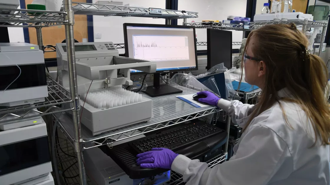A scientist in purple gloves analyzes HRMS data on a computer screen, identifying metabolites from sample injections in a well-organized lab.
