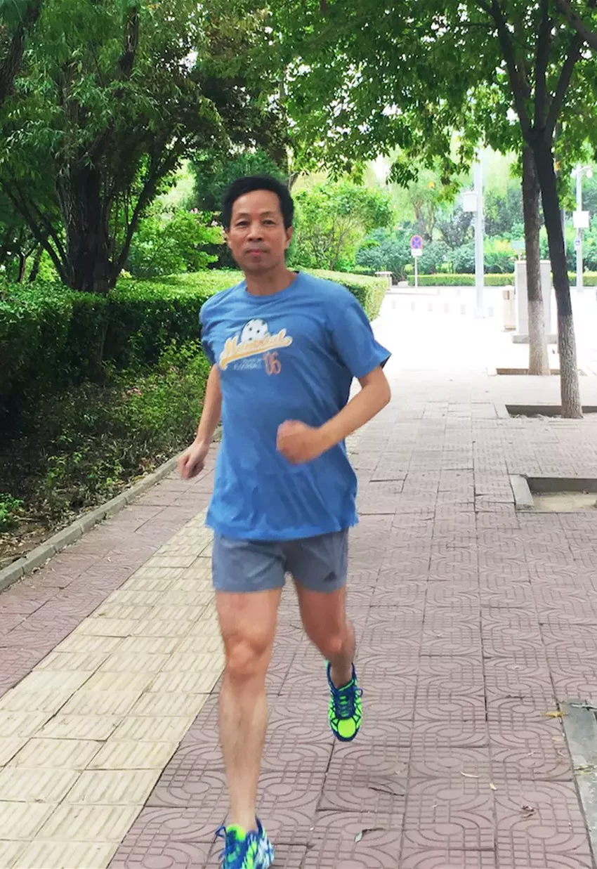 Hua-Yang - A man in a blue t-shirt and gray shorts is jogging on a tree-lined sidewalk, wearing neon running shoes, with greenery in the background.