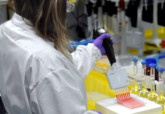A researcher wearing a white lab coat and purple gloves uses a multichannel pipette to dispense a liquid into a microplate. The laboratory workspace, focused on genetic toxicology, is filled with various scientific equipment and supplies.