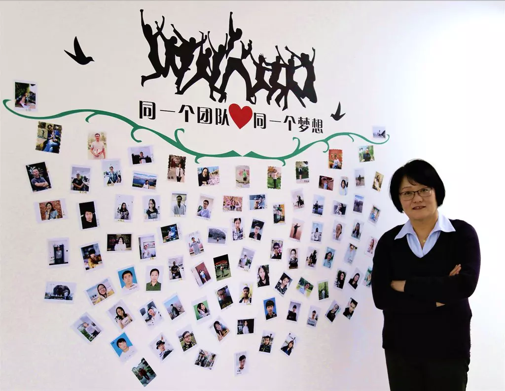 A woman stands with arms crossed beside a wall covered in small photographs, with text and a graphic of people jumping above.