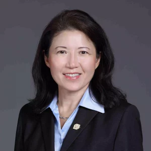 Katherine Lee, MBA - A woman with shoulder-length black hair is wearing a dark blazer over a light blue shirt, accented by a small pin on her lapel. She is smiling against a plain grey background, exuding confidence that complements her expertise in bioanalytical development.