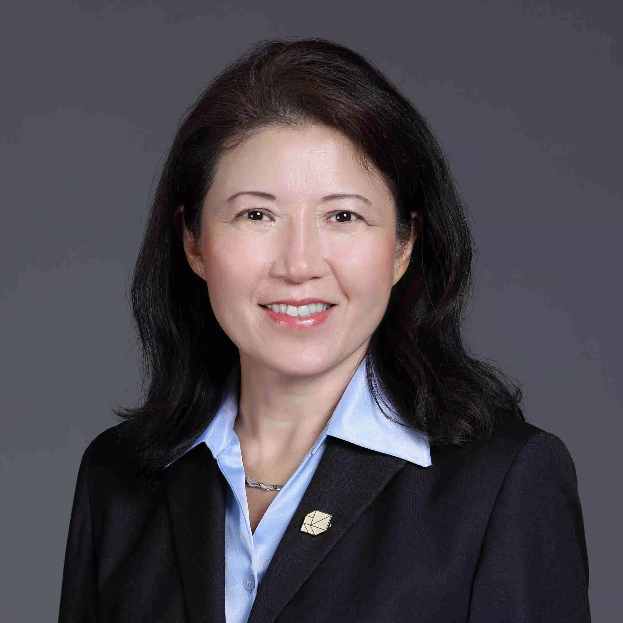 Katherine Lee, MBA - A woman with shoulder-length black hair is wearing a dark blazer over a light blue shirt, accented by a small pin on her lapel. She is smiling against a plain grey background, exuding confidence that complements her expertise in bioanalytical development.