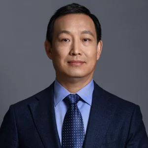 Larry Lou, EMBA, M.Eng - A man with short dark hair is seen in a professional headshot. He is wearing a dark blue suit jacket, a light blue dress shirt, and a navy blue tie with white polka dots. His arms are crossed and he is looking confidently at the camera against a plain gray background, exemplifying absolute professionalism.