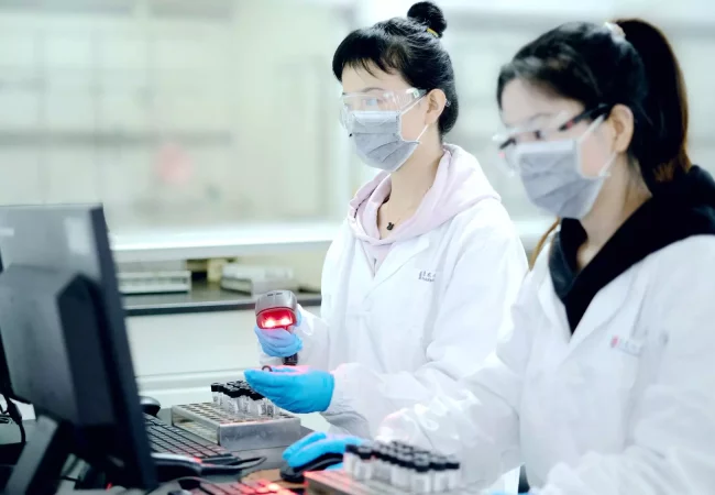 Two scientists wearing lab coats, gloves, masks, and protective goggles scan sample vials with a barcode reader, working at a laboratory computer station.