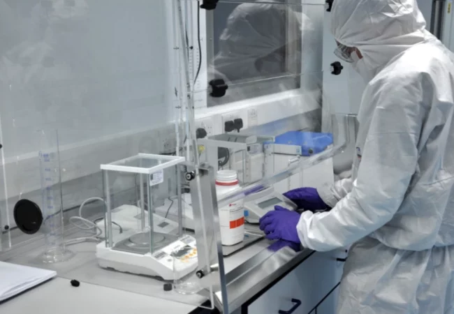 A person wearing full protective gear, including a hood, mask, gloves, and gown, works inside a laboratory. They are handling a container and working with equipment on the lab bench, which includes a scale and other scientific instruments used in preclinical ADME studies.