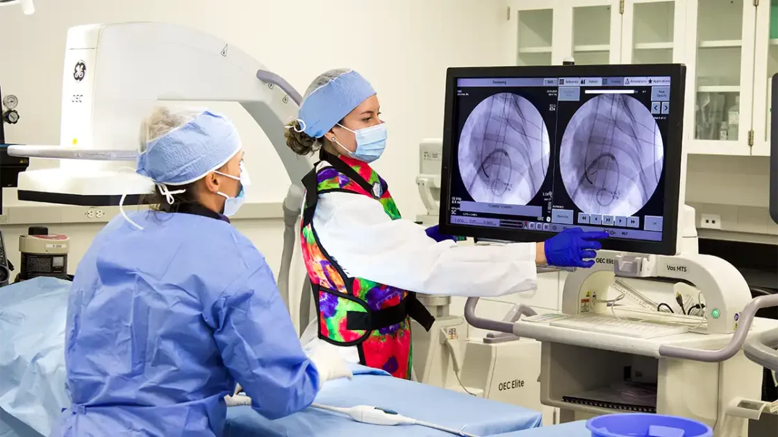 Two scientists in surgical attire conduct medical device testing using fluoroscopy imaging, reviewing real-time X-ray scans on a dual-screen monitor.