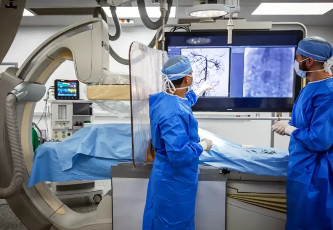 Scientists in surgical gowns review vascular imaging on monitors during advanced medical device testing in a high-tech lab environment.