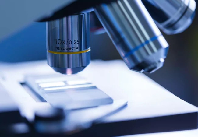 A close-up of a microscope with a 10x objective lens focuses on a slide, indicating a pathology analysis. The softly lit, sterile environment highlights detailed cellular examination.