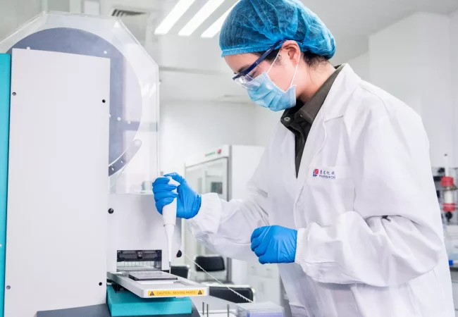 A scientist wearing a hairnet, mask, and gloves uses a pipette at a machine labeled "mosquito" in a laboratory. They are conducting in vitro disease biology studies, surrounded by lab equipment and bright overhead lighting.