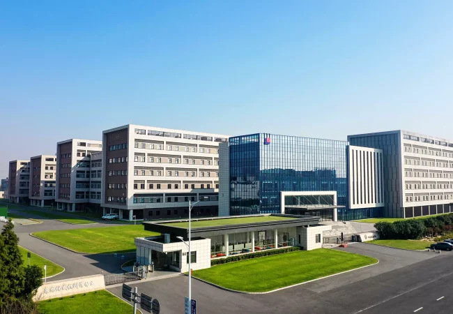 Aerial view of a large, modern office or industrial complex with multiple rectangular buildings. The central building has reflective glass windows, flanked by brick and concrete structures. This bioanalytical development hub is surrounded by well-maintained grassy areas and paved roads.