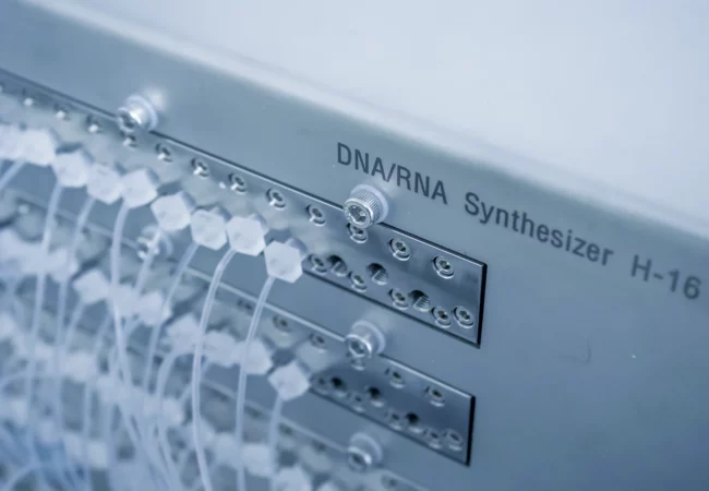 Close-up of a DNA/RNA Synthesizer H-16 machine. The device features an array of valves and white tubes connected to the front panel where it reads "DNA/RNA Synthesizer H-16." The background is slightly blurred, emphasizing the complexity of the equipment used in genetic toxicology and bioanalytical development.