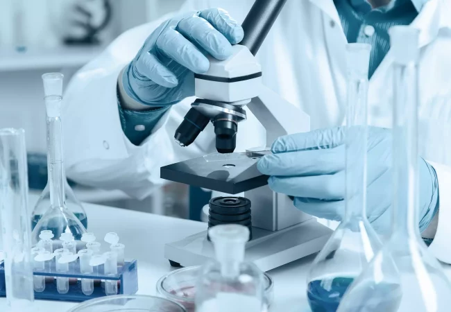 A scientist wearing gloves prepares a slide under a microscope for an ocular safety assessment. Surrounding lab glassware and vials suggest a controlled environment for detailed eye toxicity analysis.