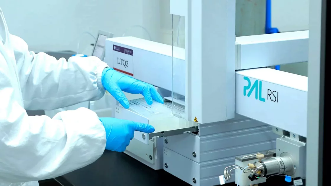 A person wearing a white lab coat and blue gloves operates a PAL RSI laboratory instrument, crucial for bioanalytical development. They handle a sample with the machine, which is situated on a black workbench in a laboratory setting. The machine has visible labeling and technical components.