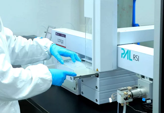 A person wearing a white lab coat and blue gloves operates a PAL RSI laboratory instrument, crucial for bioanalytical development. They handle a sample with the machine, which is situated on a black workbench in a laboratory setting. The machine has visible labeling and technical components.