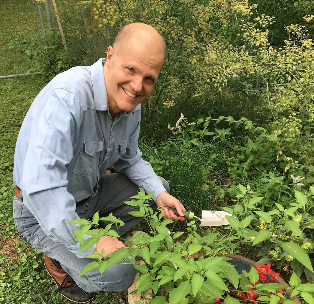 A man with a shaved head, wearing a light blue shirt, smiling while gardening outdoors, surrounded by green plants.