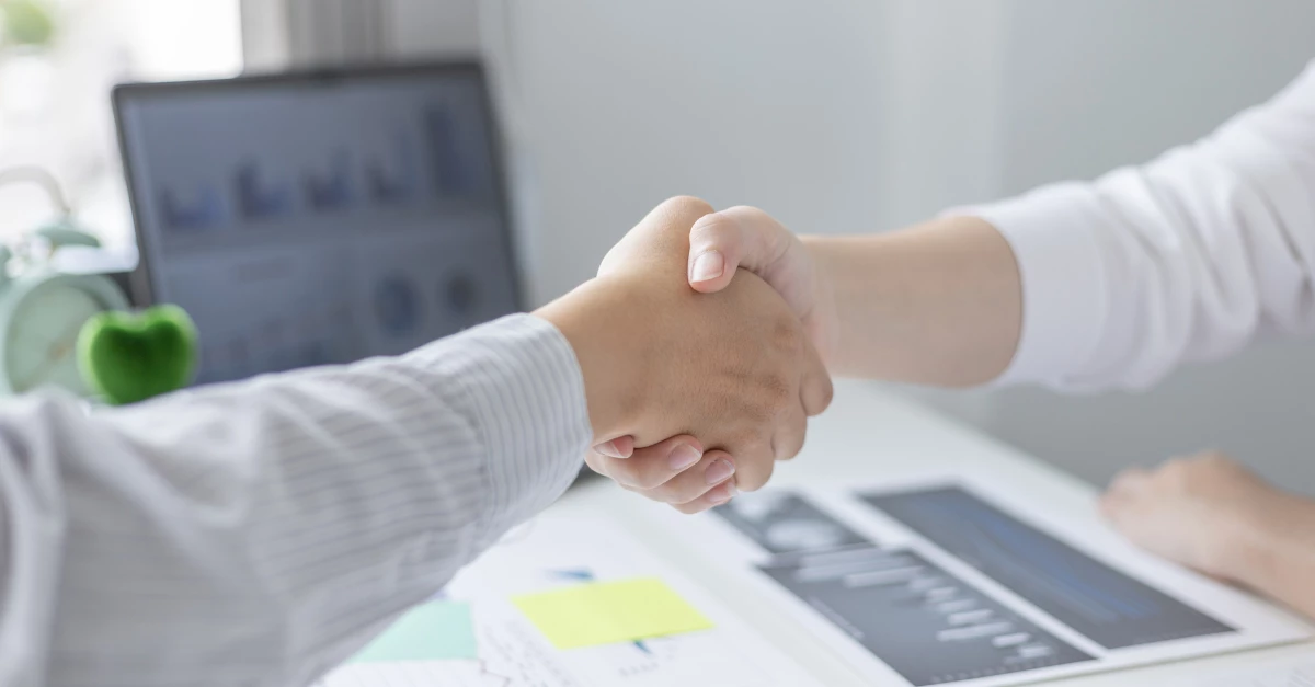 Two individuals shake hands over a desk with business charts and a laptop in the background, symbolizing partnership, collaboration, or a successful agreement. The handshake represents trust and mutual commitment in a professional setting.