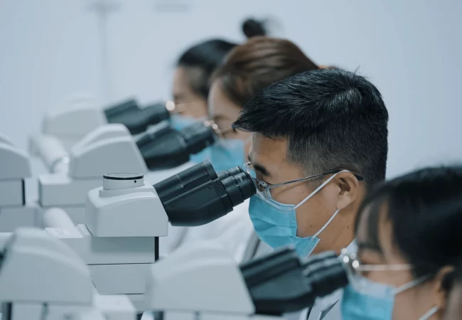 A group of scientists, wearing masks and lab coats, focus intently on microscopes in a well-lit laboratory. Their coordinated analysis suggests a collaborative pathology examination or research study.