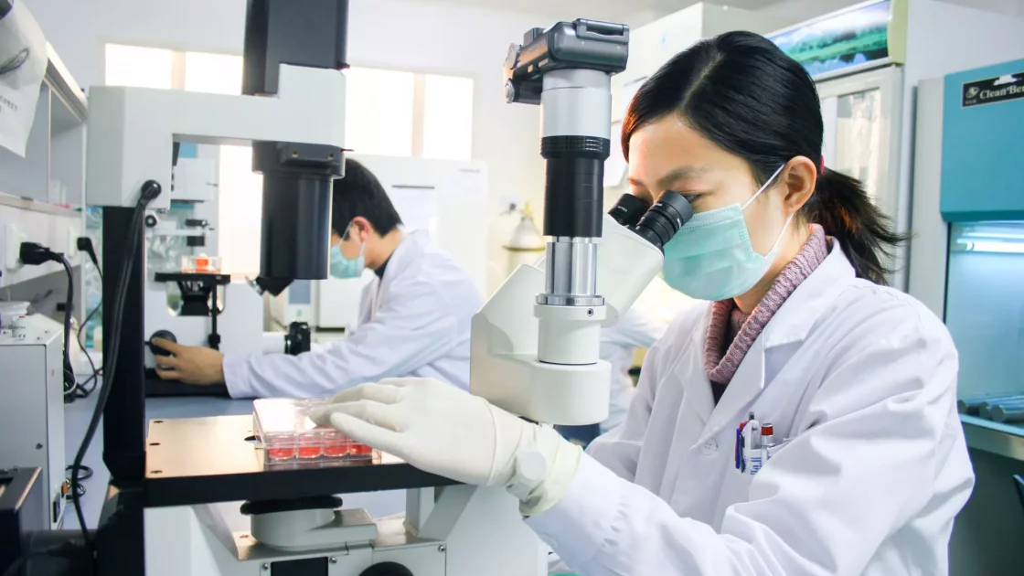 Scientists using microscopes to analyze cell samples in a laboratory, focused on pathology research with sterile equipment and protective gear.
