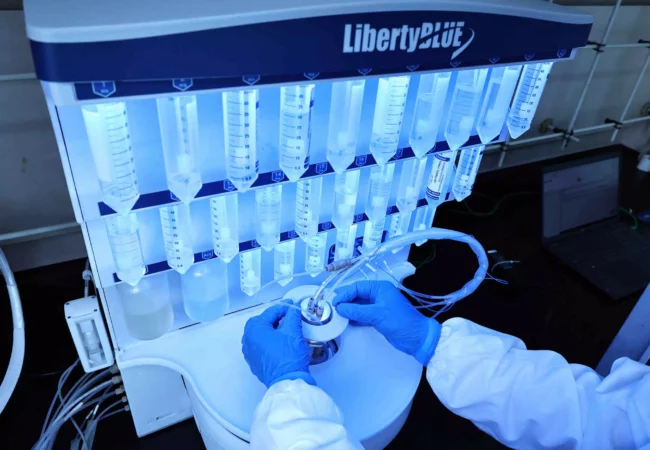 A scientist in protective gloves and a lab coat operates a Liberty Blue Automated Peptide Synthesizer, crucial for Preclinical ADME studies. The machine has multiple clear tubes with blue liquid, illuminated by blue lights. The workspace includes a laptop and various lab equipment in the background.
