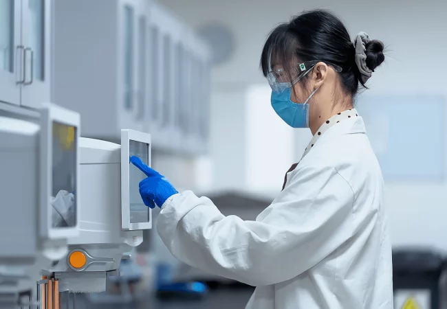 A scientist in protective gear operates lab equipment through a touchscreen, performing pharmacokinetics/pharmacodynamics (PK/PD) analysis in a controlled environment.