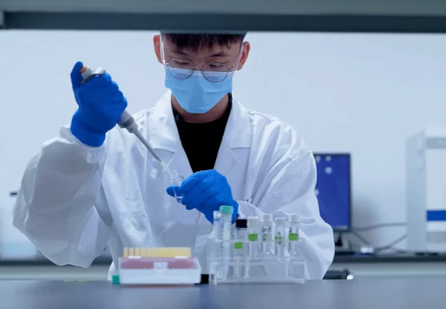 A scientist wearing gloves and a mask uses a pipette to prepare samples for pharmacokinetic/pharmacodynamic (PK/PD) analysis in a lab setting.