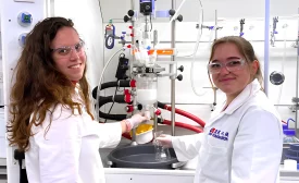 Two female scientists in lab coats and safety glasses operate a chemical reaction apparatus, smiling while working in a process chemistry lab.
