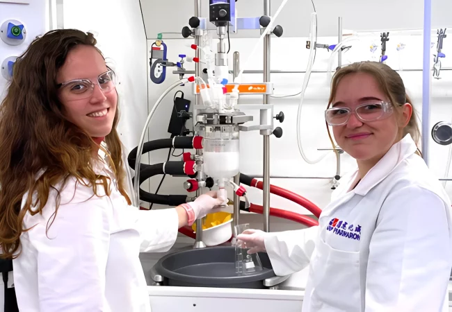Two female scientists in lab coats and safety glasses operate a chemical reaction apparatus, smiling while working in a process chemistry lab.
