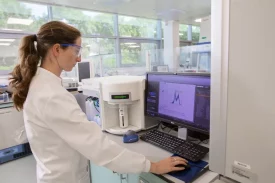 A scientist conducts product characterization and quality control (QC) analysis in a lab, working with a flow cytometer. The data on the screen displays detailed graphs and peaks, indicating an analysis of biological or pharmaceutical samples for purity, potency, and safety.