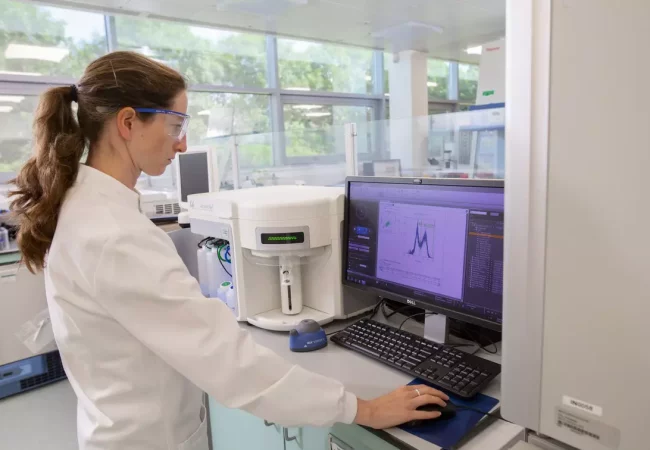 A scientist conducts product characterization and quality control (QC) analysis in a lab, working with a flow cytometer. The data on the screen displays detailed graphs and peaks, indicating an analysis of biological or pharmaceutical samples for purity, potency, and safety.
