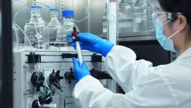 A scientist wearing a mask, safety goggles, and blue gloves is working in a laboratory. They are handling a piece of lab equipment connected to tubes and various containers, crucial for preclinical ADME studies. Bottles and glassware are visible in the background.