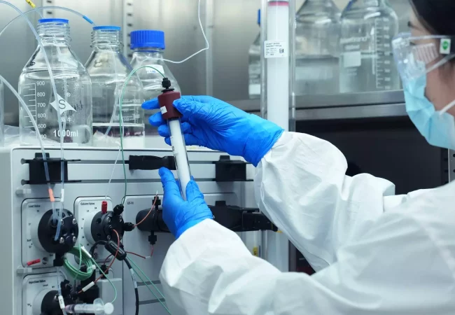 A scientist wearing a mask, safety goggles, and blue gloves is working in a laboratory. They are handling a piece of lab equipment connected to tubes and various containers, crucial for preclinical ADME studies. Bottles and glassware are visible in the background.