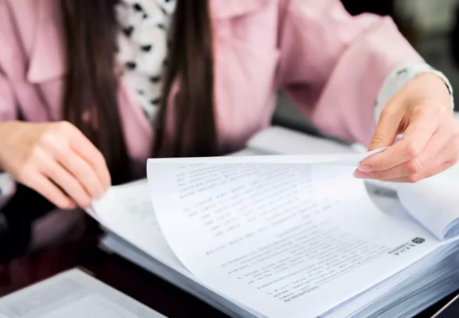 A person wearing a pink jacket and a polka-dotted shirt is seated at a desk, flipping through printed documents on preclinical ADME studies. The focus is on their hands and the papers, with no visible face. The desk has more documents and papers scattered around.