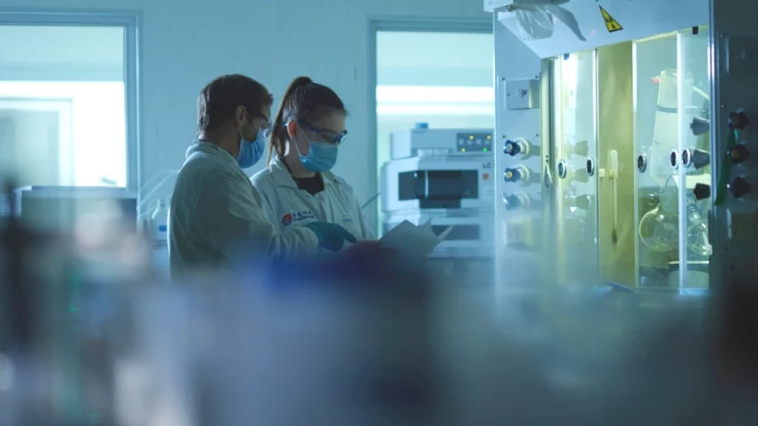 Two scientists in protective gear review documents in a lab near a radiolabelling and radiosynthesis workstation with specialized equipment.