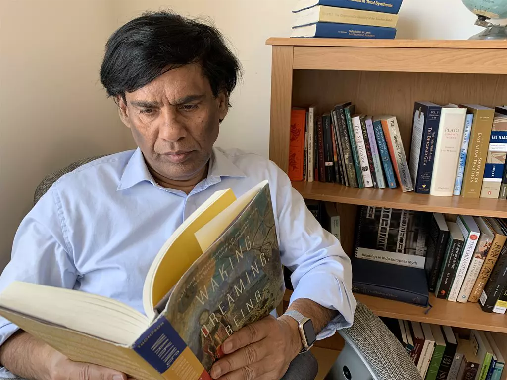 A man reading a book titled "Waking, Dreaming, Being" while sitting next to a bookshelf filled with a variety of philosophy and literature books.