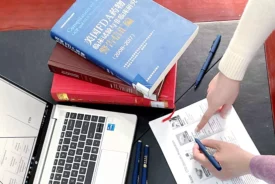 A person points to a document on a desk. Surrounding the document are a laptop, a blue pen, a black pen, and several open and closed books. The scene suggests a study or work session involving research on absolute bioavailability and note-taking for Preclinical ADME studies.
