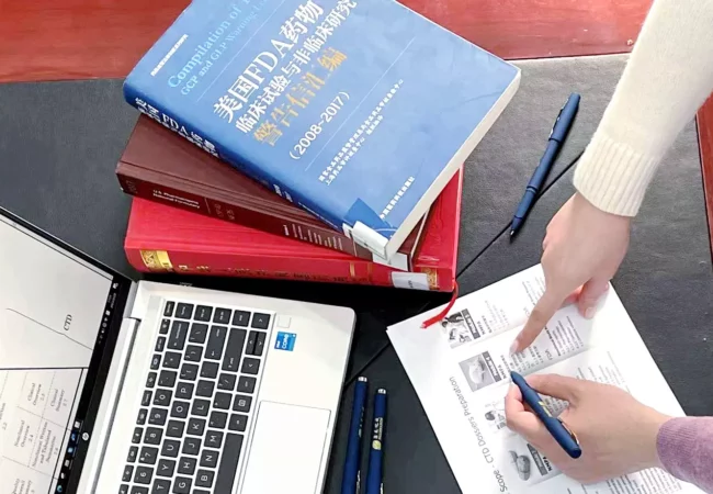 A person points to a document on a desk. Surrounding the document are a laptop, a blue pen, a black pen, and several open and closed books. The scene suggests a study or work session involving research on absolute bioavailability and note-taking for Preclinical ADME studies.