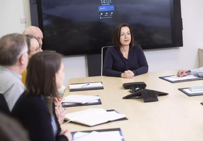A group of professionals participates in a regulatory dossier authoring meeting. The lead speaker addresses the team, surrounded by notebooks and documents, as they discuss compliance and documentation for regulatory submissions. A large screen and conference phone suggest a collaborative and formal setting.