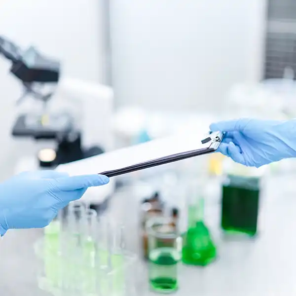 Two scientists wearing blue gloves exchange a clipboard in a lab setting with green liquid-filled glassware and a microscope in the background.