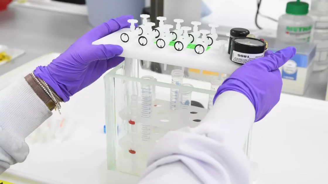 A scientist wearing purple gloves handles a filtration system with multiple syringes attached, preparing samples for environmental metabolism analysis.