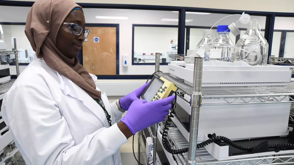 A scientist wearing a hijab, safety glasses, a white lab coat, and purple gloves is working with laboratory equipment. She holds a control panel in a lab filled with various scientific instruments and flasks, conducting preclinical ADME studies to assess genetic toxicology.