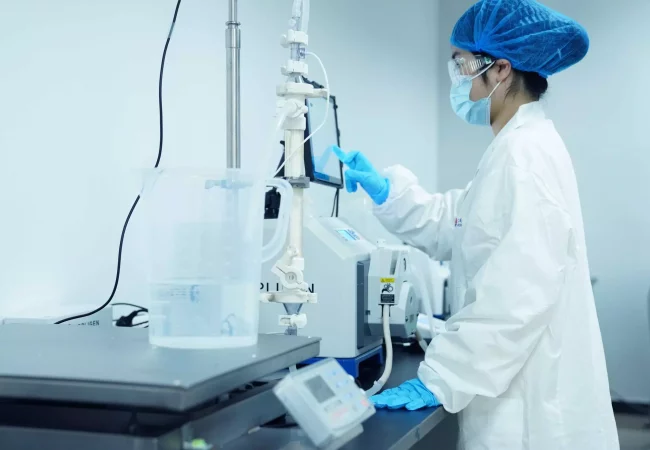 A person in a lab coat, gloves, mask, and blue hairnet operates scientific equipment in a laboratory. They are adjusting a machine connected to tubes and containers, with various lab instruments and devices on the counter nearby, contributing to bioanalytical development.