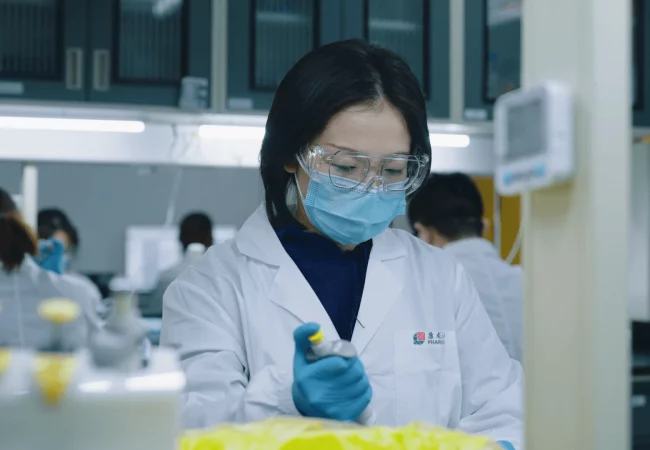 A scientist wearing protective gear, including goggles and a mask, conducts an experiment using a pipette in a lab focused on in vivo pharmacology. Other researchers work in the background, highlighting a busy research environment.