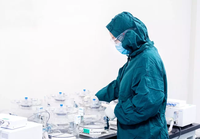 A person wearing a green protective suit, safety goggles, and a face mask works with laboratory equipment. The individual handles transparent containers connected by tubing, set on a counter in a sterile lab environment, focusing on radiolabelled ADME preclinical studies.