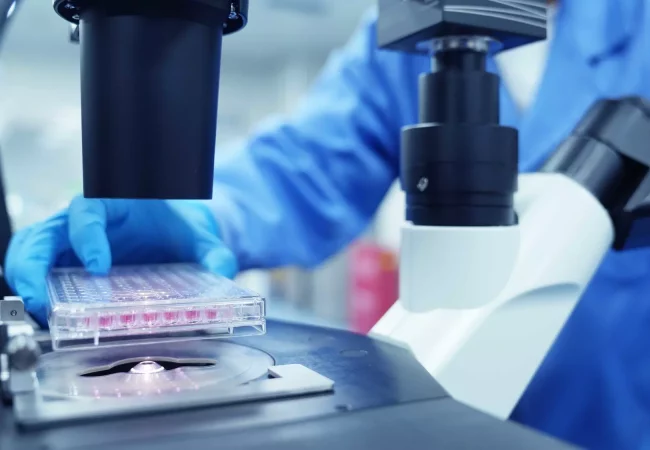 A scientist wearing blue gloves and a blue lab coat is placing a clear plastic multiwell plate under a microscope in a laboratory, conducting preclinical ADME studies. The image focuses on the microscope and the multiwell plate.