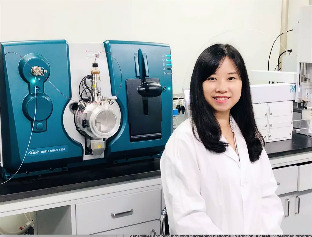 A smiling scientist in a white lab coat stands next to a SCIEX Triple Quad 5500 mass spectrometer in a laboratory setting.